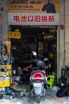 A cluttered repair shop with a motorcycle parked in front. The shop is filled with various tools, boxes, and equipment. A person is reclining on a chair inside, surrounded by spare parts and machinery. A large sign with Chinese characters is displayed above the entrance.