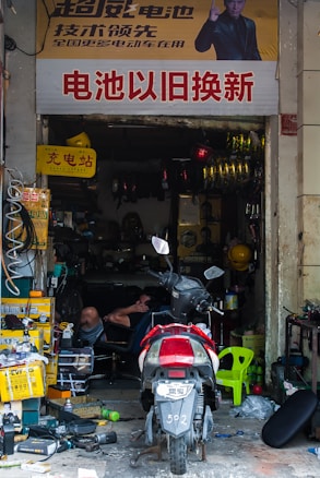 A cluttered repair shop with a motorcycle parked in front. The shop is filled with various tools, boxes, and equipment. A person is reclining on a chair inside, surrounded by spare parts and machinery. A large sign with Chinese characters is displayed above the entrance.