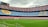 A large football stadium with brightly colored seating areas in blue and red. The seats form large patterns, including the letters and logos. The field is pristine with lush green grass, and a cloudy sky overhead adds a somber atmosphere.