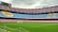 A large football stadium with brightly colored seating areas in blue and red. The seats form large patterns, including the letters and logos. The field is pristine with lush green grass, and a cloudy sky overhead adds a somber atmosphere.