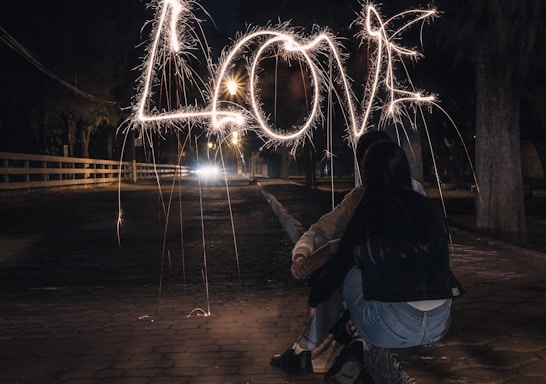 Two people are sitting closely together on a cobbled pathway at night, looking ahead at the word 'LOVE' spelled out in the air with sparklers. The atmosphere is intimate and illuminated by street lamps in the background.