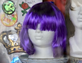A mannequin head displays a vibrant purple wig with bangs covering the eyes. The background contains an assortment of items including a crown emblem, a floral pattern featuring a rose, and a cartoon character badge.