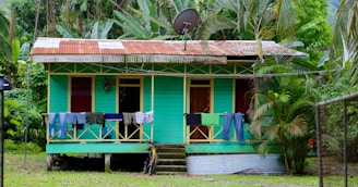A small wooden house with a teal exterior is surrounded by lush tropical plants and trees. The roof is made of corrugated metal, and it has a satellite dish on top. Clothes hang to dry on the porch railing, and stairs lead up to the entrance. A fence and a bicycle are visible in the foreground.