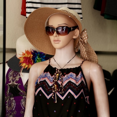 A mannequin dressed in a patterned black dress with colorful geometric designs. It wears large sunglasses and a wide-brimmed straw hat adorned with a polka dot bow. Behind it, another hat with a colorful floral decoration is visible, along with various clothing items.