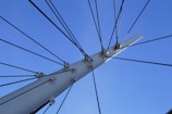 A collection of black cables radiates outwards from a white, metallic pole or structure, set against a clear blue sky. The cables are fastened with metal hardware, creating a symmetrical and organized appearance.