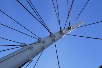 A collection of black cables radiates outwards from a white, metallic pole or structure, set against a clear blue sky. The cables are fastened with metal hardware, creating a symmetrical and organized appearance.
