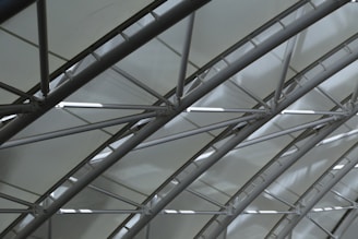 A series of curved metal beams create a geometric pattern against a translucent ceiling, allowing diffused light to filter through. The structure exudes a modern and industrial aesthetic, with a focus on the interplay of lines and shadows.