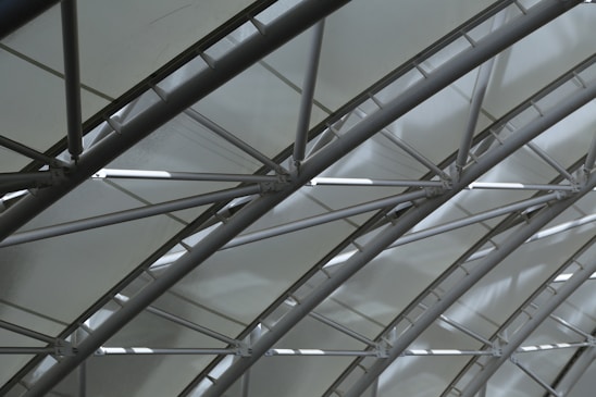 A series of curved metal beams create a geometric pattern against a translucent ceiling, allowing diffused light to filter through. The structure exudes a modern and industrial aesthetic, with a focus on the interplay of lines and shadows.