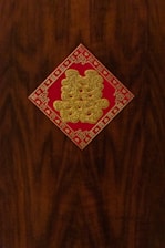 A decorative, red square with gold glitter Chinese characters is affixed to a dark wooden surface. Intricate patterns and symbols frame the edges of the square.
