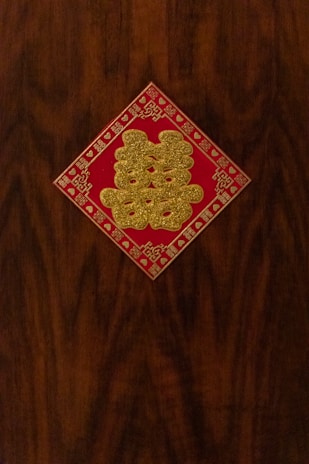 A decorative, red square with gold glitter Chinese characters is affixed to a dark wooden surface. Intricate patterns and symbols frame the edges of the square.