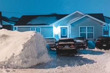 A suburban house is partially obscured by a large pile of snow with several cars parked in the driveway. The area is covered in snow, creating a wintery scene. The house is light beige with a dark shingle roof, illuminated by the blue-tinted light near the entrance.