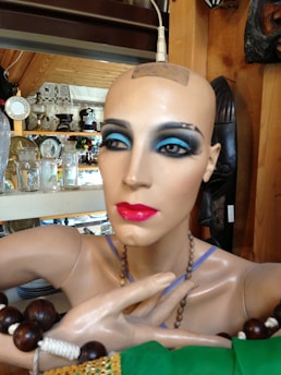 A mannequin with bold makeup, featuring bright blue eyeshadow and vivid red lipstick. It is adorned with various accessories, such as beaded necklaces and bracelets. The mannequin is set against a backdrop of wooden shelves displaying assorted glassware and decorative items.