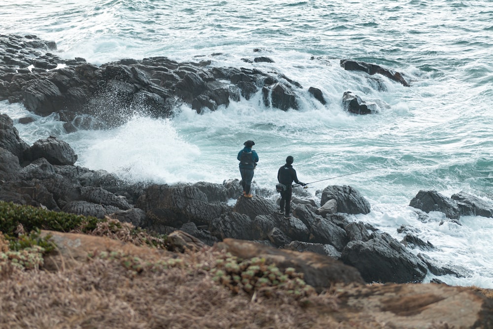 昼間の海の波の近くの岩の上に立っている黒いジャケットの男