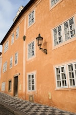 A well-preserved orange stucco building with multiple white-framed windows and a black vintage-style street lamp on its side. The building has an inclined cobblestone sidewalk in front and a historic plaque indicating the year 1685. The architecture features a combination of simple rectangular shapes and traditional design elements.