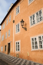 A well-preserved orange stucco building with multiple white-framed windows and a black vintage-style street lamp on its side. The building has an inclined cobblestone sidewalk in front and a historic plaque indicating the year 1685. The architecture features a combination of simple rectangular shapes and traditional design elements.