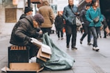 An elderly individual is sitting on the street with a stack of cardboard boxes and a large green tarp. Passersby wearing winter clothing walk past without paying attention. The scene suggests an urban environment, possibly during a colder season.