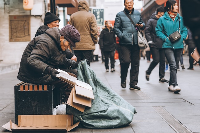 An elderly individual is sitting on the street with a stack of cardboard boxes and a large green tarp. Passersby wearing winter clothing walk past without paying attention. The scene suggests an urban environment, possibly during a colder season.