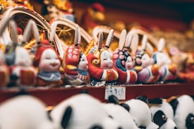 A collection of colorful, ornamental rabbit figures in traditional attire are neatly arranged in rows. The focus is on the intricate details and vibrant colors of the figures, which have distinct facial expressions. Soft lighting provides a warm atmosphere, highlighting the craftsmanship.