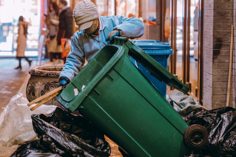 child in brown knit cap and brown jacket sitting on green plastic trash bin