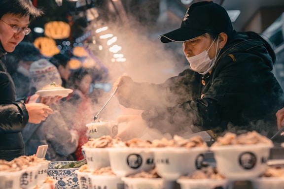 A vendor wearing a mask and cap is serving steaming hot food from a pot into a bowl in an indoor market setting. Another person is holding a plate of food, with lights and a busy atmosphere in the background.