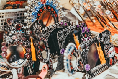 A collection of decorative handheld mirrors with intricate designs and embellishments, featuring jewels, vibrant tassels, and intricate patterns. The scene includes multiple ornate mirrors displayed together, alongside other ornate objects.