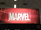 A red and black sign prominently displaying the word 'MARVEL' in bold white capital letters, set within a white rectangular border. The background features a subtle pattern of comic-style artwork, creating a dynamic and engaging visual.