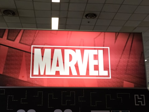 A red and black sign prominently displaying the word 'MARVEL' in bold white capital letters, set within a white rectangular border. The background features a subtle pattern of comic-style artwork, creating a dynamic and engaging visual.