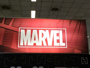 A red and black sign prominently displaying the word 'MARVEL' in bold white capital letters, set within a white rectangular border. The background features a subtle pattern of comic-style artwork, creating a dynamic and engaging visual.