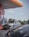 A gas station features prominently with a close-up view of a sleek, dark-colored car in the foreground. In the background, a silver SUV is positioned near the fuel pumps, with trees and a clear blue sky visible. The station displays characteristic branding and signage, creating a modern and everyday setting.