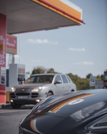A gas station features prominently with a close-up view of a sleek, dark-colored car in the foreground. In the background, a silver SUV is positioned near the fuel pumps, with trees and a clear blue sky visible. The station displays characteristic branding and signage, creating a modern and everyday setting.