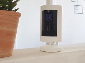 A white security camera is positioned on a white surface, with a terracotta potted plant visible on the left side of the image. The blurred background contains framed pictures on a white wall.