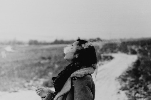 A person stands outdoors in a winter landscape, exhaling a cloud of smoke into the cold air. The ground is covered with a light dusting of snow, and the background is a blurred, rural landscape. The person is wearing a coat with a fur collar, suggesting cold weather.