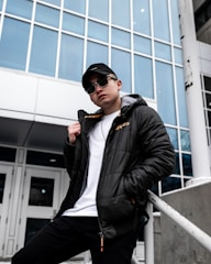 A person in a black jacket, white shirt, and black cap leans against a white railing in front of a modern building with large glass windows. The individual is wearing sunglasses and has a casual pose, suggesting confidence.