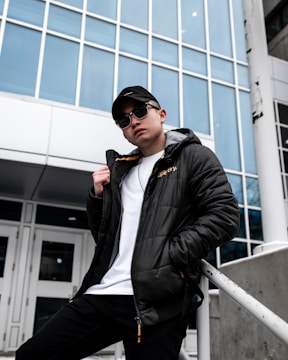 A person in a black jacket, white shirt, and black cap leans against a white railing in front of a modern building with large glass windows. The individual is wearing sunglasses and has a casual pose, suggesting confidence.