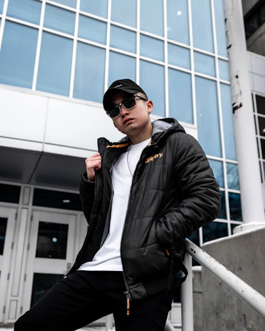 A confident person wearing a tailored jacket and casual pants, leaning against a modern building wall.