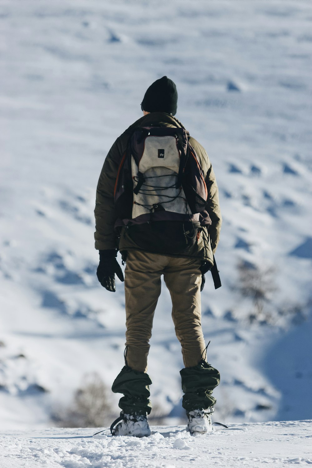 Hombre en chaqueta negra y pantalones marrones con mochila negra caminando sobre suelo cubierto de nieve durante