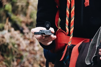 A person is holding a compass in their hand, with a background of blurred greenery. They are wearing black and red outdoor clothing with a colorful striped scarf.