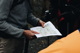 Several individuals are gathered, focusing on a map held by one person. The person holding the map is wearing a gray jacket and carrying a backpack, indicative of a hiking or outdoor setting.