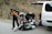 A group of people is preparing for an outdoor adventure next to a white van with the back door open. They are organizing camping gear and clothing on the ground, surrounded by bare trees on a quiet road. One person is adjusting their jacket while others gather equipment.
