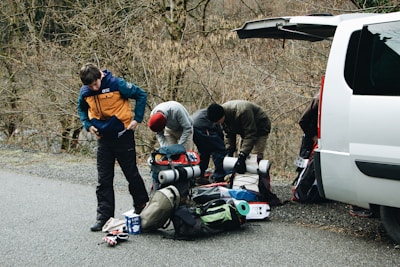 A group of people is preparing for an outdoor adventure next to a white van with the back door open. They are organizing camping gear and clothing on the ground, surrounded by bare trees on a quiet road. One person is adjusting their jacket while others gather equipment.