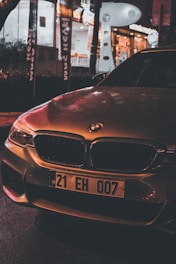 A BMW car parked on a wet street at night, illuminated by the warm glow of streetlights and nearby shop signs. The car's license plate is visible, displaying '21 EH 007'. In the background, there are various illuminated signs and storefronts, suggesting a lively urban setting.