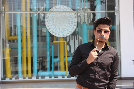 A person wearing sunglasses and a black shirt holds a stick with a fake mustache in front of their face. The background features a glass window with a decorative display of vertical yellow and blue pipes, along with a circular design containing text.