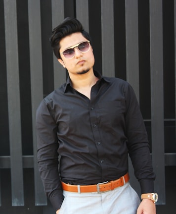 Man wearing a sleek black shirt and gray pants standing against a minimalist urban backdrop.