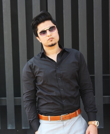 A man is wearing a black button-up shirt, light gray pants, and a tan belt. He accessorizes with sunglasses and a wristwatch. His posture is relaxed, with one hand in his pocket, and he stands against a vertical striped black backdrop.