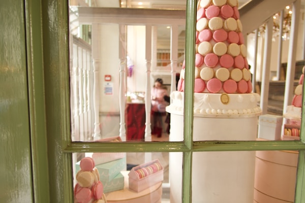 An elegantly arranged display of pink and cream-colored macarons forms a tower, situated near a window with a green frame. Pastel-colored macaron boxes are visible at the base of the tower, surrounded by soft lighting that adds a warm glow to the setting.