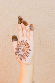An ornate henna design is applied to the palm of an upturned hand. The fingers are partially covered with a thick layer of henna paste. The intricate pattern on the palm features circular and floral elements with fine detailing. A textured, circular background provides contrast to the lighter skin tone of the hand.