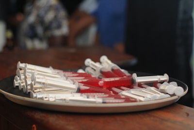 A plate on a wooden surface is filled with multiple syringes, each containing a red liquid. The background is blurred, with indistinct figures and colors, focusing attention on the plate of syringes in the foreground.
