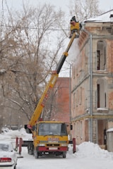 A yellow crane vehicle is positioned next to a building, extending its arm to a worker in a basket who is clearing snow from the roof. The building appears old, with worn walls, and there are leafless trees in the background. The ground and rooftops are covered with snow, indicating a winter setting.