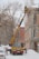 A yellow crane vehicle is positioned next to a building, extending its arm to a worker in a basket who is clearing snow from the roof. The building appears old, with worn walls, and there are leafless trees in the background. The ground and rooftops are covered with snow, indicating a winter setting.
