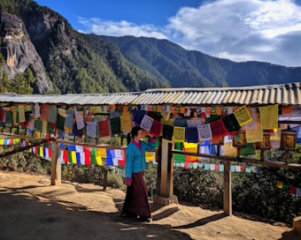 A person in traditional attire stands near a structure adorned with colorful Tibetan prayer flags, surrounded by a scenic mountainous landscape. The clear blue sky and green forested hills provide a tranquil backdrop.